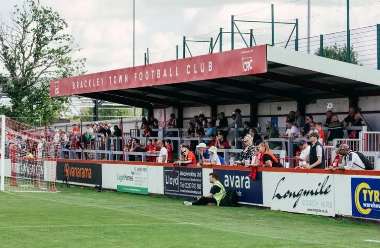 Bầu không khí sôi động tại sân St. James Park Brackley vào một ngày thi đấu, khán giả đứng gần sân tạo nên sự gần gũi đặc trưng của các trận đấu bóng đá hạng thấp ở Anh.